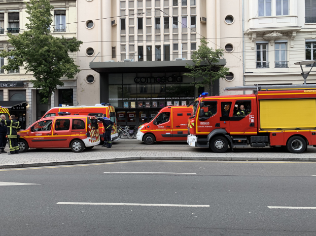 Eine Gruppe von Feuerwehrautos auf einer Straße in Paris geparkt, mit Menschen auf dem nächsten Gehweg, Gebäuden, Bäumen und einem Fahrrad im Hintergrund.