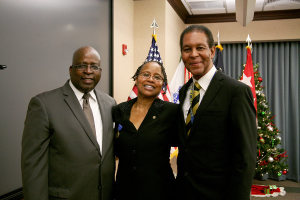 Eine Gruppe strahlender Menschen vor einem geschmückten Weihnachtsbaum und einem Vorhang, mit Flaggen und verschiedenen Gegenständen im Hintergrund und einem Tuch mit Gegenständen auf der rechten Seite.