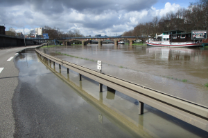 Eine überflutete Straße mit einem Boot im Wasser, eine Brücke im Hintergrund, Bäume, Gebäude, Pfosten, Bretter und ein bewölkter Himmel.