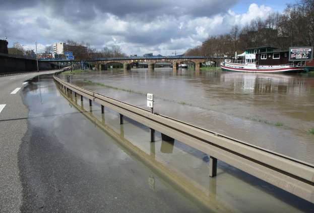 Eine überflutete Straße mit einem Boot im Wasser, eine Brücke im Hintergrund, Bäume, Gebäude, Pfosten, Bretter und ein bewölkter Himmel.