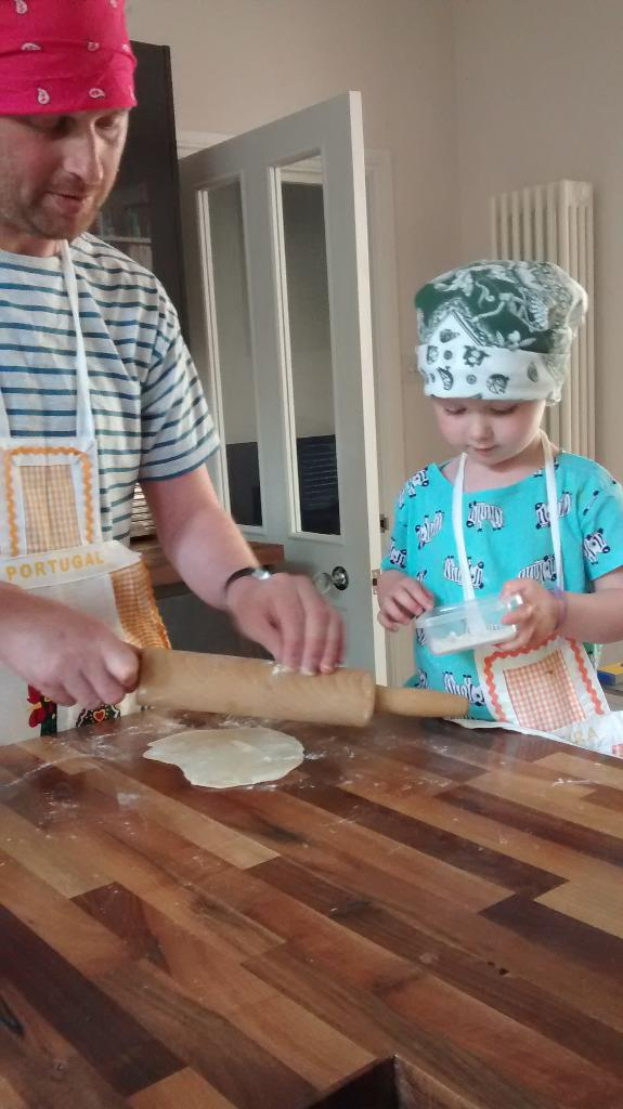 Ein Mann und ein Kind, beide mit Schürzen und Mützen bekleidet, stehen vor einer Tür mit Griff; der Mann hält eine Rolle, während das Kind einen Behälter hält, mit Essen auf dem Tisch dazwischen.