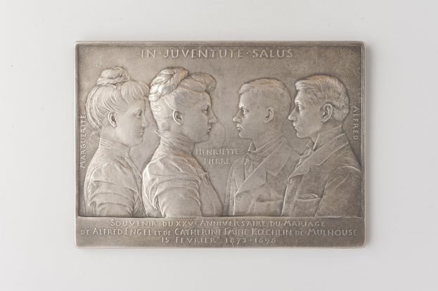 Silberne Plakette mit einem eingravierten Bild von drei Personen und einem Gedenktext auf einem weißen Hintergrund.