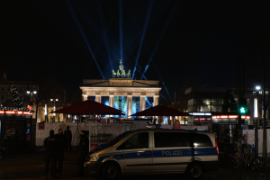 Ein Polizeifahrzeug parkt nachts vor dem Brandenburger Tor in Berlin, umgeben von ein paar Menschen und diversen Gegenständen wie Schirmen, Gebäuden, Bäumen, Lichtern, Masten und einer Ampel.