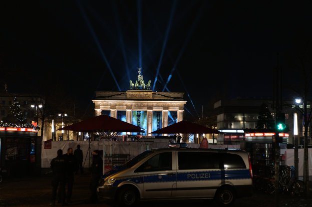 Ein Polizeifahrzeug parkt nachts vor dem Brandenburger Tor in Berlin, umgeben von ein paar Menschen und diversen Gegenständen wie Schirmen, Gebäuden, Bäumen, Lichtern, Masten und einer Ampel.