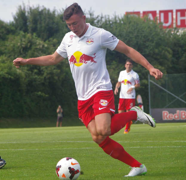 Professioneller Fußballspieler, der einen Ball auf einem Rasenfeld kickt, mit "RB Leipzig"-Schriftzug im Hintergrund bei klarem Himmel.