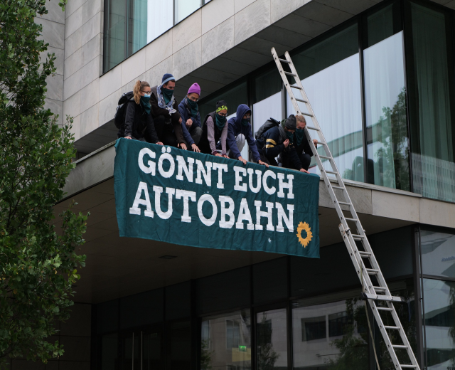 Gruppe von Menschen auf einem Gebäudedach mit einem Banner, mit einem Baum links und Glasfenstern im Hintergrund, wahrscheinlich protestierend für die deutsche Regierung.