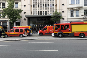 Eine Gruppe von Feuerwehrautos auf einer Straße in Paris geparkt, mit Menschen auf dem nächsten Gehweg, Gebäuden, Bäumen und einem Fahrrad im Hintergrund.