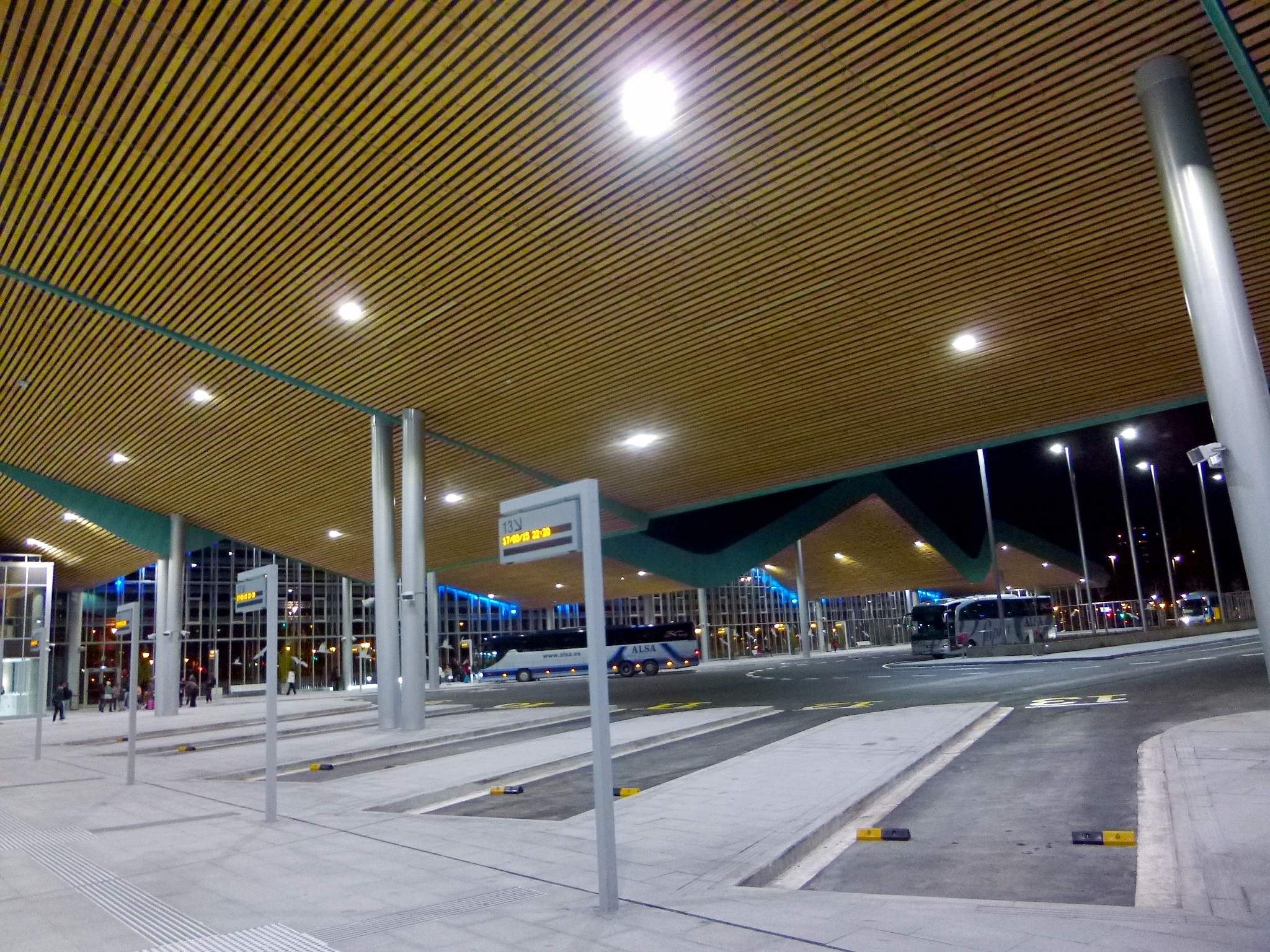 Bus-Haltestelle bei Nacht mit Deckenbeleuchtung, Fahrzeugen auf der Straße, Beschilderung, Menschen und beleuchtete Gebäude im Hintergrund.