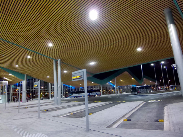 Bus-Haltestelle bei Nacht mit Deckenbeleuchtung, Fahrzeugen auf der Straße, Beschilderung, Menschen und beleuchtete Gebäude im Hintergrund.