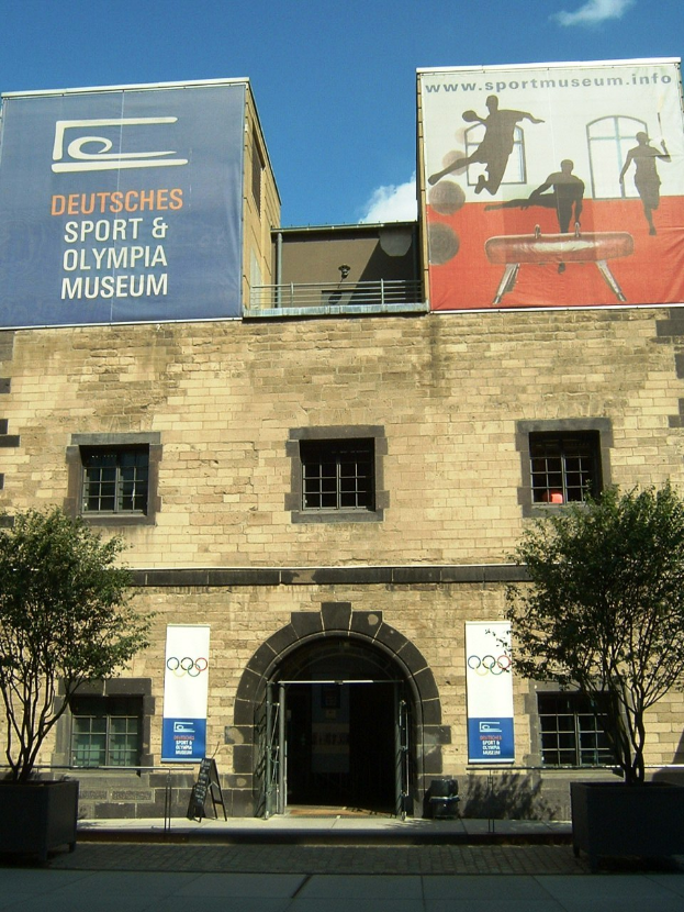 Außenansicht des Deutschen Sport & Olympia Museums in Berlin mit einem Gebäude mit Fenstern und einer Tür, umgeben von Bäumen und Bannern mit Text und Bildern, unter einem bewölkten Himmel.