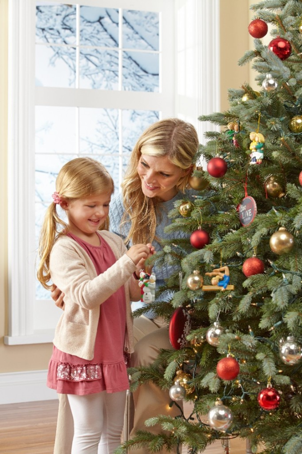 Ein geschmückter Weihnachtsbaum steht in einem Haus mit einer Frau und einem Kind im Vordergrund, einem Fenster und einer Wand im Hintergrund und Bäumenoutside des Fensters.