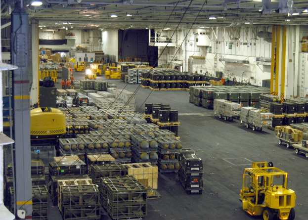 Großes Lagerhausinnere mit mehreren Lastwagen, Containern, Menschen, Deckenbeleuchtung, Metallstangen und einem sichtbaren Rad.