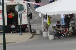 Ein Krankenwagen steht auf der linken Seite einer Straße, während auf der rechten Seite ein Zelt viele Menschen schützt, mit einem Tisch und einem Stuhl in der Nähe.