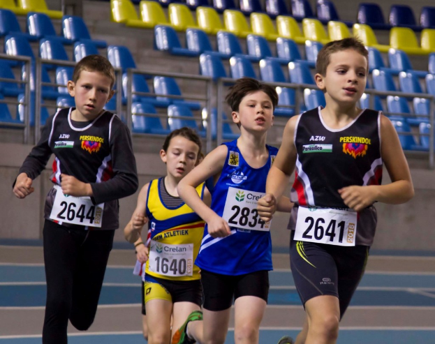 Vier Personen rennen, jede trägt ein Kleid in einer anderen Farbe mit einem Aufkleber, blaue und gelbe Stühle sind im Hintergrund sichtbar.