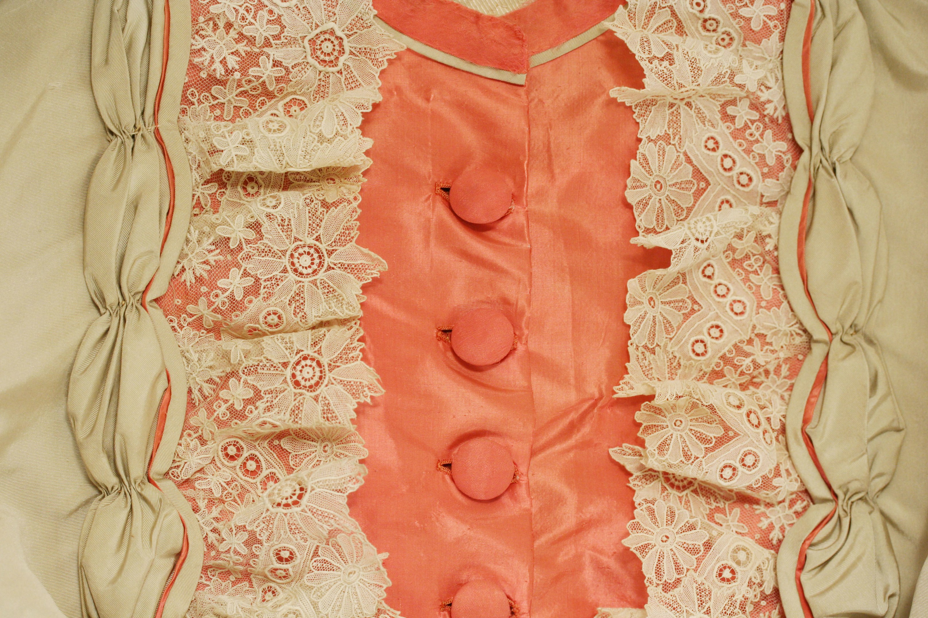 Nahaufnahme eines rosafarbenen und cremefarbenen Kleides mit weißer Spitze und Knöpfen vorne, das einen klassischen und eleganten Stil zeigt.