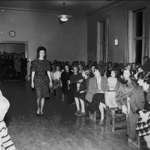 Eine Frau in einem schwarzen und weißen Kleid läuft über den Laufsteg, während das Publikum bei einer Modenschau zur Feier des Frauenwahlrechts zuschaut.