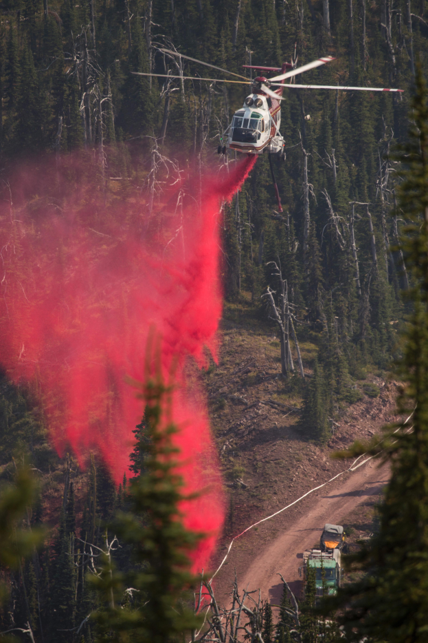 Ein Hubschrauber fliegt und wirft Wasser auf einen Waldbrand, mit einem Fahrzeug auf der rechten Seite und vielen Bäumen im Hintergrund.