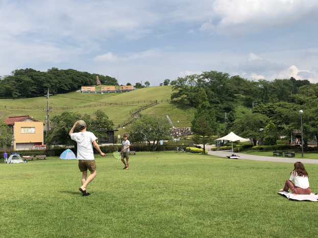 Gruppe von Menschen, die Badminton in einem Park spielen, mit einem Mann, der einen Schläger hält, und einer Frau, die auf einem Tuch auf dem Gras sitzt, vor dem Hintergrund von Zelten, Straßenmasten, Straßenlaternen, Strommasten, Stromkabeln, Gebäuden, Bäumen, Hügeln und einem bewölkten Himmel.