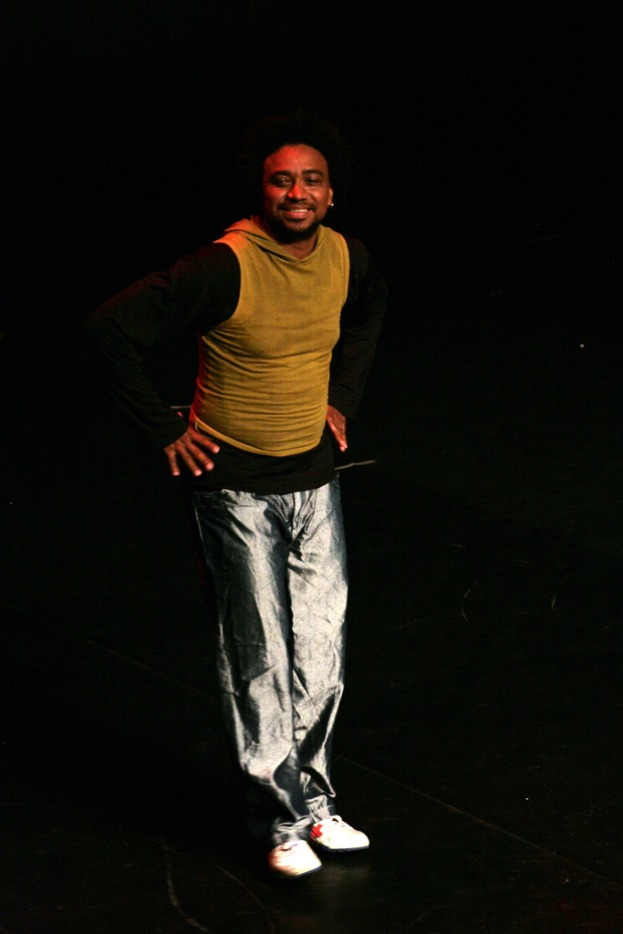 Eine Person in einem T-Shirt lächelt und performs auf einer Bühne mit dunklem Hintergrund.