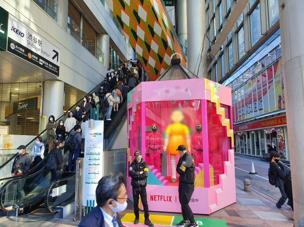 Eine Gruppe von Menschen steht vor einem Gebäude mit Glaswänden, Säulen und Texttafeln, einige tragen Masken, mit einer Rolltreppe im Blick; eine pinkfarbene Box in der Mitte zeigt Schaufensterpuppen und trägt 'Netflix' neuestes Netflix-Booth in Tokyo'.