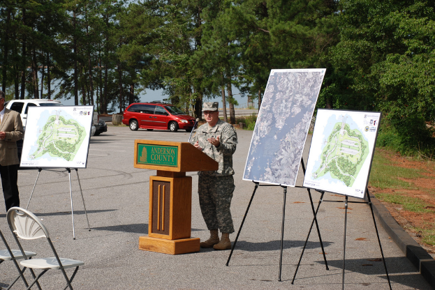 Eine Person steht an einem Rednerpult mit Mikrofon und schaut auf Plakate, Stühle, entfernte Fahrzeuge und Bäume.