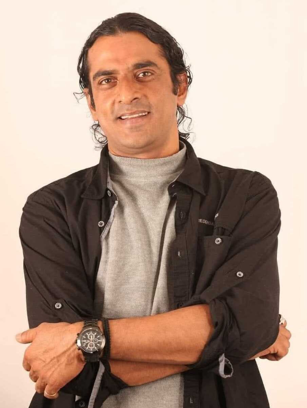 Ein Mann in einem schwarzen und aschfarbenen Outfit mit überkreuzten Armen, der eine Uhr trägt und vor einem weißen Hintergrund steht, der in Gedanken versunken zu sein scheint.