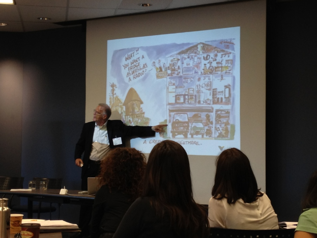 Mann in weißem Hemd und schwarzem Blazer zeigt auf ein Fahrzeug auf einem Bildschirm in einem Konferenzsaal, mit drei Frauen im Vordergrund und einem Tisch mit einer Wasserflasche, einem Glas und einem Buch links.
