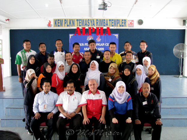 Eine Menschenmenge, die fröhlich posiert und vor einer Bühne fotografiert wird, mit einer Wandtafel im Hintergrund, einem Tischventilator rechts, einem Podium links und einem Projektor oben im Bild.