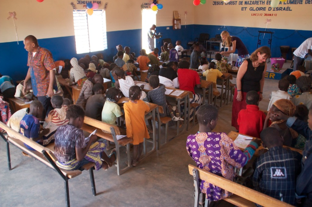 Kinder auf Bänken im Vordergrund, mit Erwachsenen, die sie führen, und im Hintergrund Stühle, eine Tür, ein Fenster und ein Tisch, sowie Ballons und Text oben.