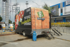 Ein Lastwagen vor einem Gebäude mit einer Tafel, auf der "Teilen Sie eine positiv überladene Erfahrung" steht, mit Stufen, einem Zylinder auf dem Boden, Geländern, Strommasten mit Drähten, Laternenpfählen, Bäumen und einem bewölkten Himmel im Hintergrund.