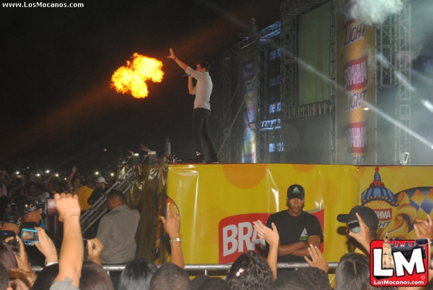 Eine Gruppe von Menschen, einige mit Kameras, ein Mann in der Mitte vor einem Feuer, umgeben von Metallstangen, Werbetafeln und Rauch, mit einem Wasserzeichen oben links und einem Logo unten rechts.