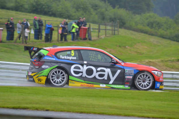 Ein Auto auf einer Rennstrecke, das Rauch ausstößt, umgeben von Gras, einem Zaun, Mülltonnen, Menschen, Bäumen und einem klaren blauen Himmel.
