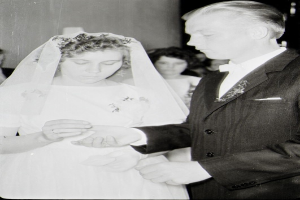 Ein Schwarz-Weiß-Foto einer Braut in einem weißen Kleid und Schleier und eines Bräutigams in einem Anzug, die ihre Eheringe tauschen, mit ein paar Menschen im Hintergrund.