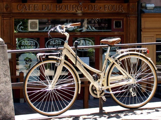 Ein weißes Fahrrad steht auf einem Fußweg mit einem Rohrgeländer dahinter und einem Holz- und Glashaus im Hintergrund.