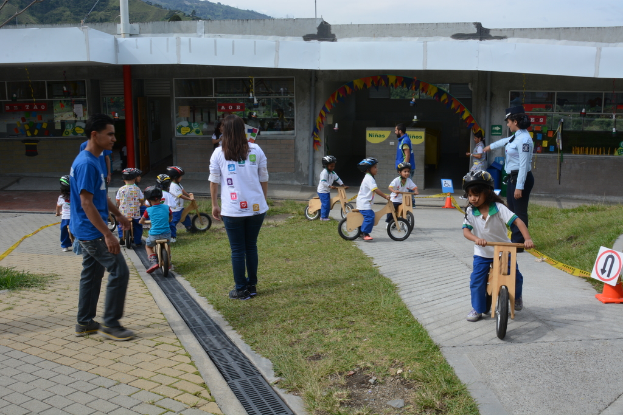 Kinder mit Helmen fahren Fahrräder auf einer grünen Wiese, Erwachsene stehen in der Nähe und ein Schild und ein Gebäude sind im Hintergrund zu sehen.