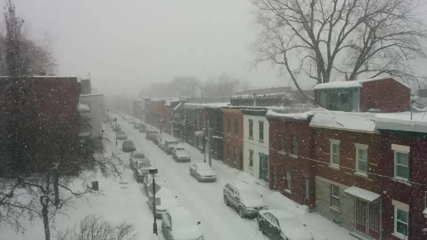 Eine schneebedeckte Straße mit Autos und Gebäuden auf beiden Seiten und Bäumen im Hintergrund.