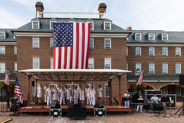 Eine Gruppe von Menschen auf einer Bühne in Mount Vernon, einige spielen Musikinstrumente, mit Flaggen, Lautsprechern, Pflanzen und einem bewölkten Himmel im Hintergrund.