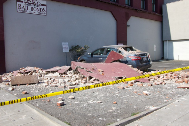 Eine zerbrochene Ziegelwand liegt auf dem Boden mit verstreuten roten Ziegeln, ein geparktes Auto, eine Topfpflanze und ein Gebäude mit einer "Kautionskaution"-Plakette im Hintergrund.