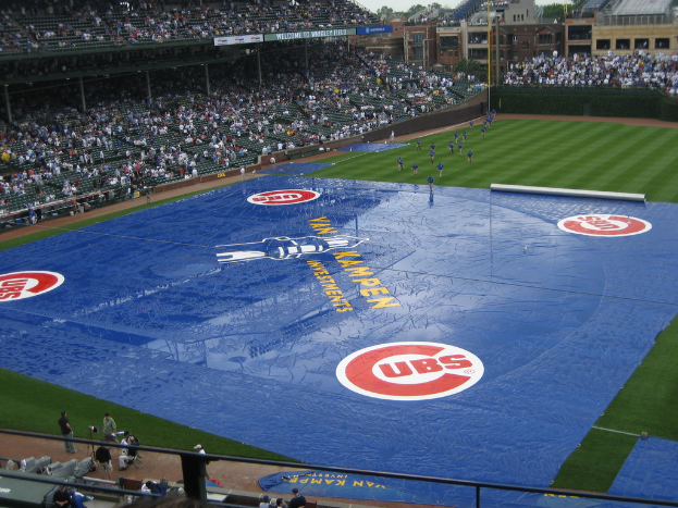 Eine Stadion-Szene mit Menschen, die auf dem Boden spielen und andere auf Stühlen sitzen, Gebäude, Fenster, Bäume und Himmel im Hintergrund sowie Schilder mit Text, eine Kamera-Stative und andere Gegenstände auf dem Boden.