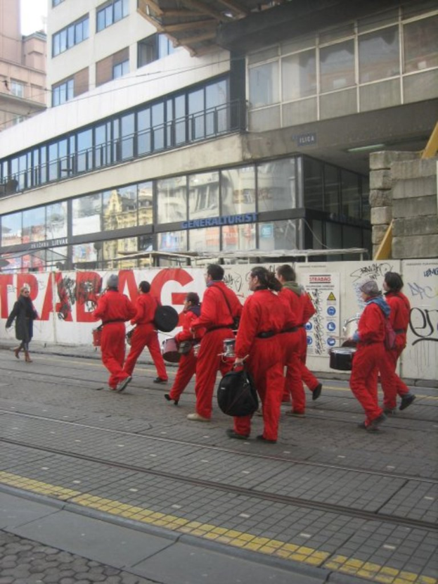 Menschen, die auf einem Boden gehen und Musikinstrumente spielen, mit einer Umzäunungswand im Hintergrund, die Plakate und Text trägt, und weiter hinten Glaswand- und Fenstergebäude mit Namen darauf.