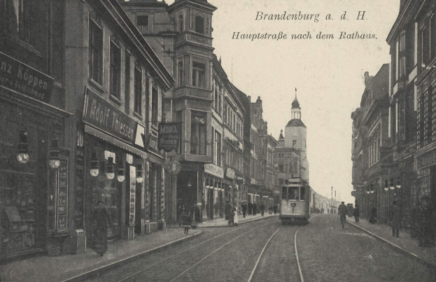 Altes Schwarz-Weiß-Foto einer Straßenkreuzung in Brandenburg, Deutschland mit einer Tram, Fußgängern, Gebäuden, Schildern, Straßenlaternen und einem bewölkten Himmel.