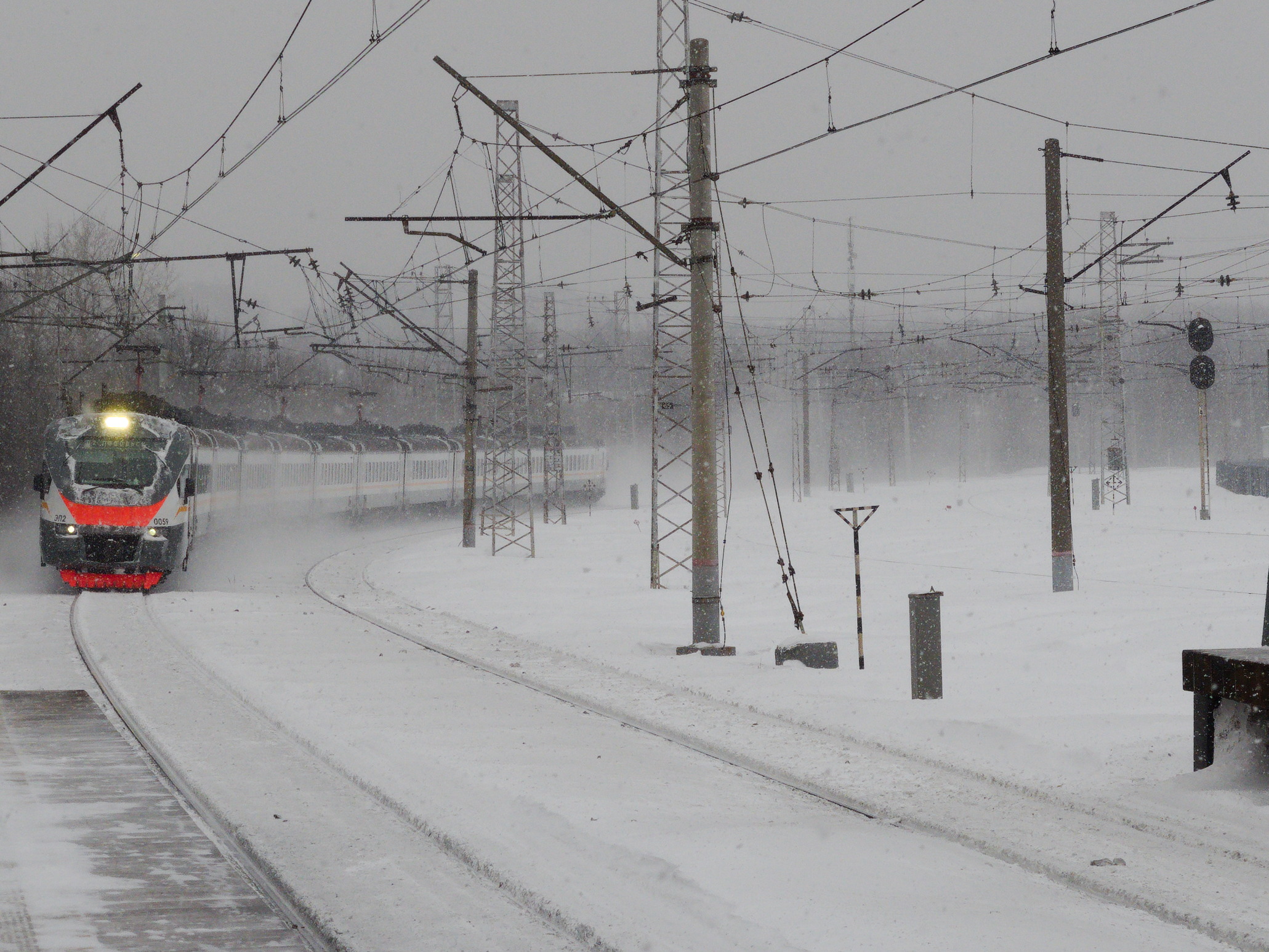 Ein Zug fährt auf schneebedeckten Schienen mit Strommasten und Drähten daneben, eine Bank auf der rechten Seite und Bäume mit einem klaren Himmel im Hintergrund.