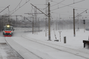 Ein Zug fährt auf schneebedeckten Schienen mit Strommasten und Drähten daneben, eine Bank auf der rechten Seite und Bäume mit einem klaren Himmel im Hintergrund.