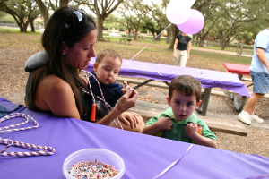 Eine Frau in Schutzbrille hält ein Baby, daneben steht ein Junge in einem grünen Kleid, neben Tischen mit einer Geigenpartitur und einer Schale mit Würfeln, Ballons und einem Mann rechts im Bild, mit Bäumen im Hintergrund.