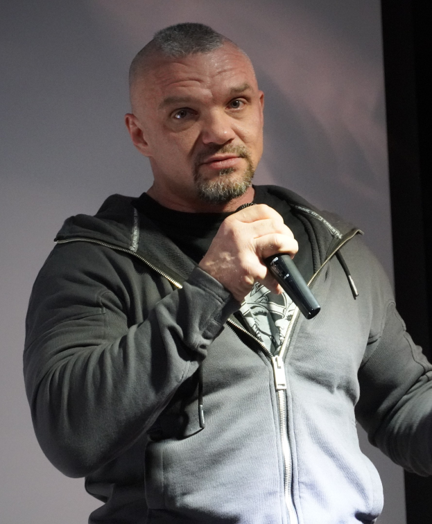 Ein Mann in einer schwarzen Kapuzenjacke und einem schwarzen Anzug mit einem wei├čen Hemd, der ein Mikrofon haltend, selbstbewusst vor einer Wand steht und wie ein Komiker wirkt.