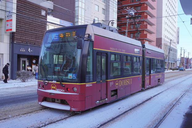 Ein roter und gelber Zug fährt eine verschneite Straße entlang, gesäumt von hohen Gebäuden, Strommästen, Schildern, Pflanzen und einer Gruppe von Menschen unter einem bewölktem Himmel.