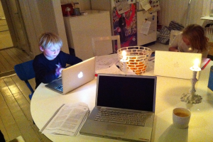Ein Studiertisch mit zwei Kindern, einem Jungen mit einem Laptop und einem Mädchen, das arbeitet, mit drei Laptops, einem Buch, einer Tasse Tee, zwei brennenden Kerzen, einem Fenster, einem weiteren Tisch und einem Kühlschrank.