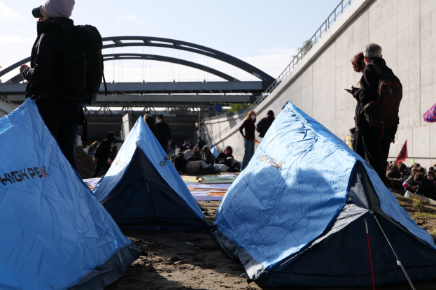 Gruppe von Menschen, die auf einem sandigen Strand in der Nähe von Zelten sitzen, mit einer Wand und einer Brücke im Hintergrund, die an einer Klimawandeldemonstration teilnehmen.