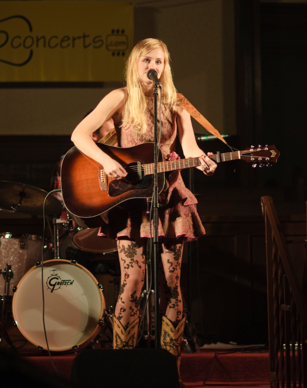 Eine Frau, die auf der BÃ¼hne eine Gitarre spielt und singt, mit einer Band im Hintergrund und einem Bildschirm an der Wand dahinter.