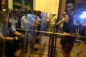Eine Frau in einem schwarzen T-Shirt und roten Shorts steht mit einem Mikrofon in der Hand vor einer Reihe von Einsatzpolizisten hinter einer Metallabsperrung, umgeben von Zuschauern und Gebäuden im Hintergrund.